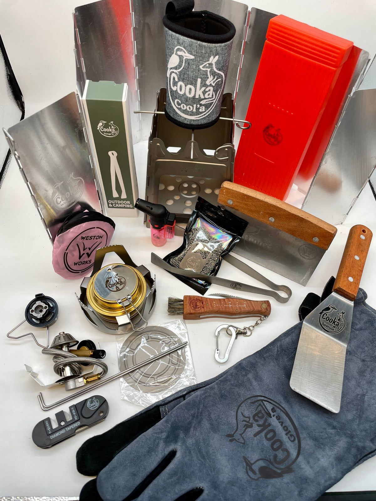 Cooka Big Kahuna Kit laid out: titanium folding stove, Burn'a metho stove, butane adaptor, heat-resistant glove, tongs, spatula, Cut'a knife and Sharpen'a